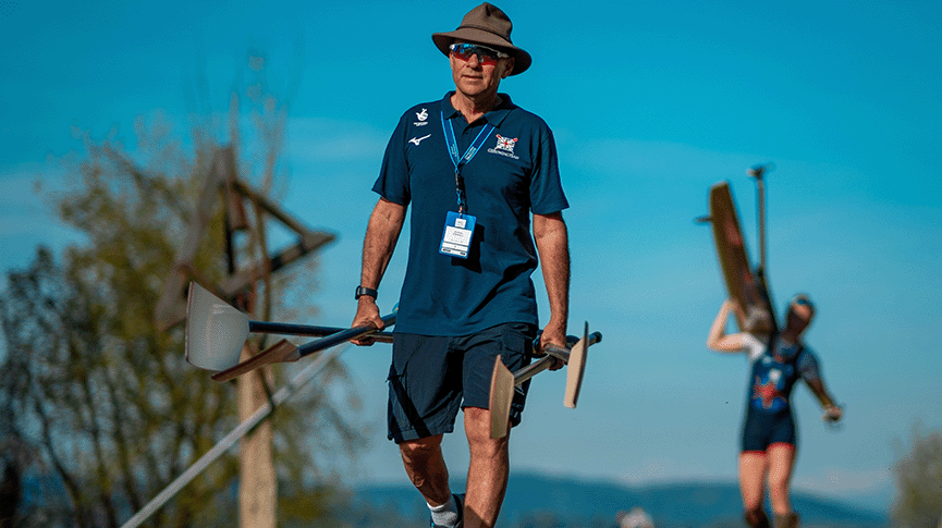 Male coach carrying four sculling blades with single sculler carrying boat in the background